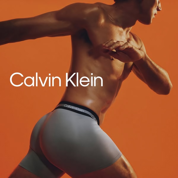 A high-energy, close-up shot of a male athlete in gray boxer briefs against a bold orange backdrop, emphasizing product fit and athletic physique.